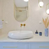 Elegancka łazienka z białą umywalką, złotymi bateriami i lustrem. Ściana za umywalką wyłożona jest mozaiką w jodełkę. Półka z płytek w kabinie prysznicowej jest dyskretnie widoczna w odbiciu lustra.