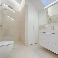 Nowoczesna łazienka z podwieszaną toaletą i minimalistyczną umywalką. Ściany i podłoga wyłożone jasnymi płytkami, które tworzą spójną, elegancką całość.