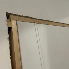 Widoczna szpara między ościeżnicą a ścianą, odsłaniająca drewnianą konstrukcję i fragment białych drzwi.