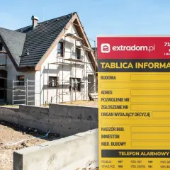 Tablica informacyjna budowy domu w trakcie prac, z widocznym rusztowaniem i fundamentami.