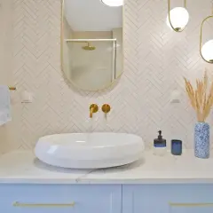 Elegancka łazienka z białą umywalką, złotymi bateriami i lustrem. Ściana za umywalką wyłożona jest mozaiką w jodełkę. Półka z płytek w kabinie prysznicowej jest dyskretnie widoczna w odbiciu lustra.