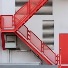 Czerwone, metalowe schody zewnętrzne prowadzą do czerwonych drzwi. Widać kratki wentylacyjne i instalacje. Idealne, by pokazać, jak zabudować schody zewnętrzne.