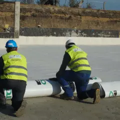 Dwóch pracowników w kamizelkach odblaskowych przygotowuje hydroizolację. Rozwijają rolki materiału, by zabezpieczyć teren budowy przed wilgocią.