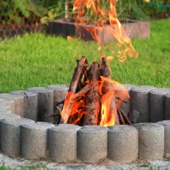 Płonące ognisko przy domu, otoczone kamiennym kręgiem, na tle zielonej trawy.