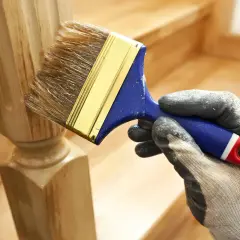 Ręka w rękawicy nakłada lakier na drewniany słupek schodów. Dowiedz się, jak odnowić stare schody drewniane.
