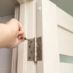 Dłoń dokręca śrubę mocowania zawiasu w ościeżnicy białych drzwi.