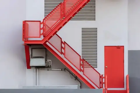 Czerwone, metalowe schody zewnętrzne prowadzą do czerwonych drzwi. Widać kratki wentylacyjne i instalacje. Idealne, by pokazać, jak zabudować schody zewnętrzne.
