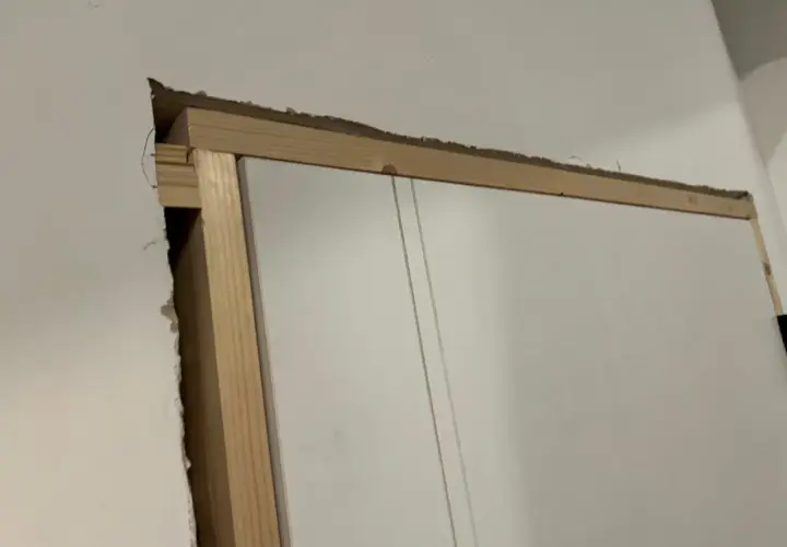 Widoczna szpara między ościeżnicą a ścianą, odsłaniająca drewnianą konstrukcję i fragment białych drzwi.