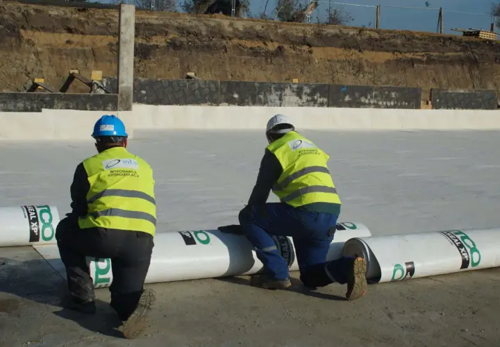 Dwóch pracowników w kamizelkach odblaskowych przygotowuje hydroizolację. Rozwijają rolki materiału, by zabezpieczyć teren budowy przed wilgocią.