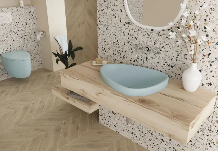 Jasna łazienka z błękitną misą umywalki, drewnianym blatem i podłogą w jodełkę. W tle błękitna miska WC i terazzo. Boczne światło podkreśla faktury.
