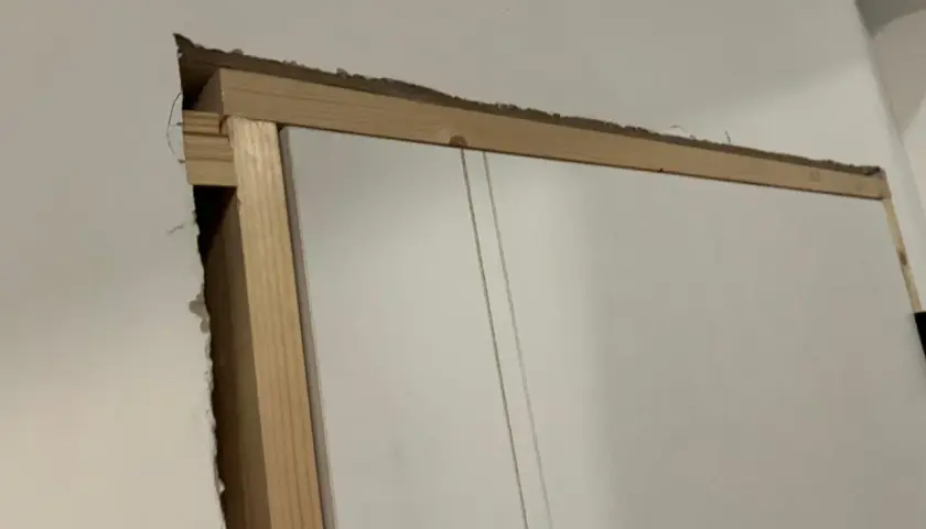 Widoczna szpara między ościeżnicą a ścianą, odsłaniająca drewnianą konstrukcję i fragment białych drzwi.