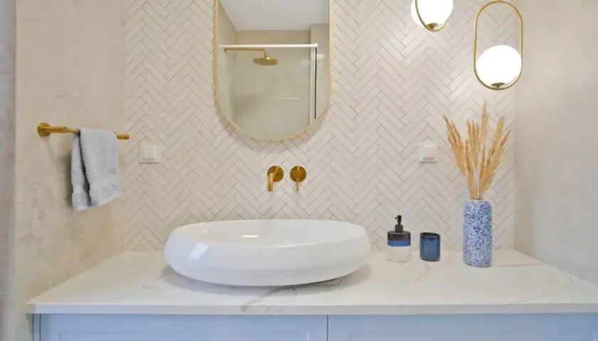 Elegancka łazienka z białą umywalką, złotymi bateriami i lustrem. Ściana za umywalką wyłożona jest mozaiką w jodełkę. Półka z płytek w kabinie prysznicowej jest dyskretnie widoczna w odbiciu lustra.