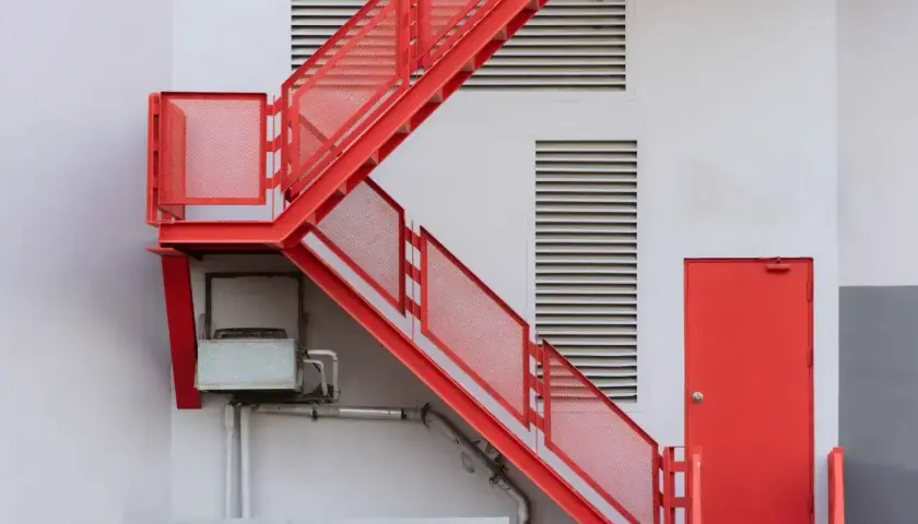 Czerwone, metalowe schody zewnętrzne prowadzą do czerwonych drzwi. Widać kratki wentylacyjne i instalacje. Idealne, by pokazać, jak zabudować schody zewnętrzne.