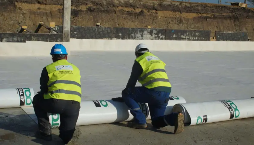 Dwóch pracowników w kamizelkach odblaskowych przygotowuje hydroizolację. Rozwijają rolki materiału, by zabezpieczyć teren budowy przed wilgocią.