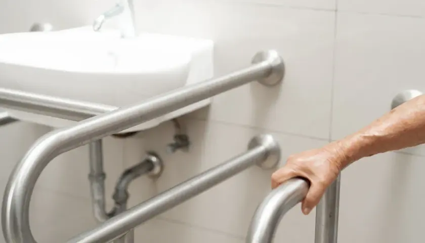 Dłoń osoby starszej chwyta uchwyt dla niepełnosprawnych. Łazienka przystosowana zgodnie z przepisami.