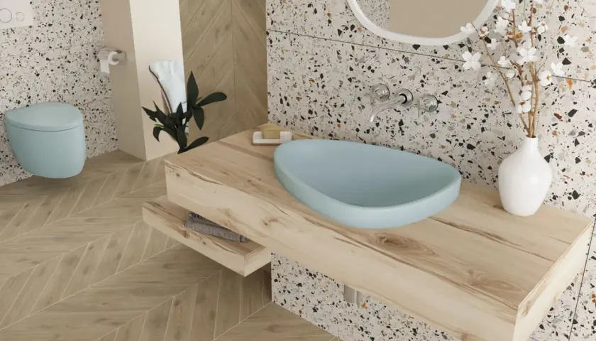 Jasna łazienka z błękitną misą umywalki, drewnianym blatem i podłogą w jodełkę. W tle błękitna miska WC i terazzo. Boczne światło podkreśla faktury.
