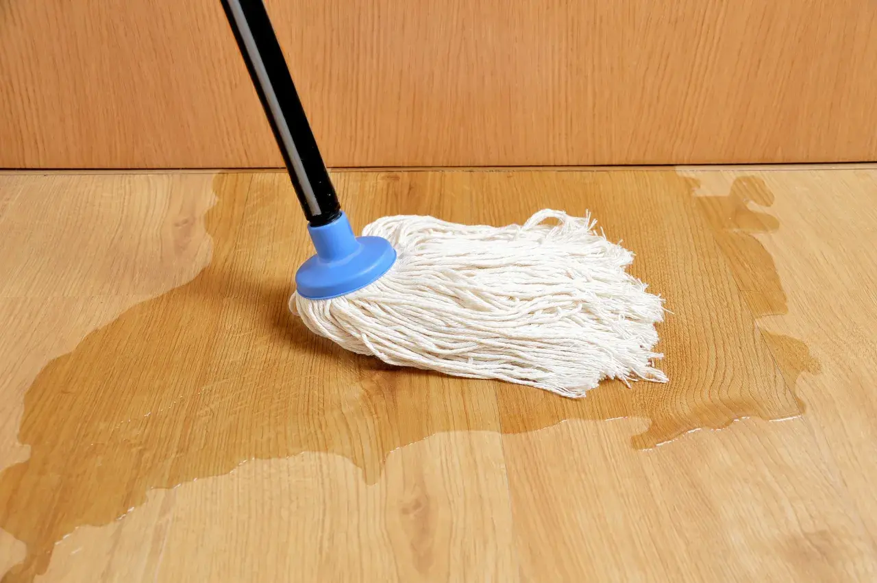 Mop na drewnianej podłodze, gdzie widać zalane panele na łączeniach. Woda rozlewa się po jasnym drewnie.
