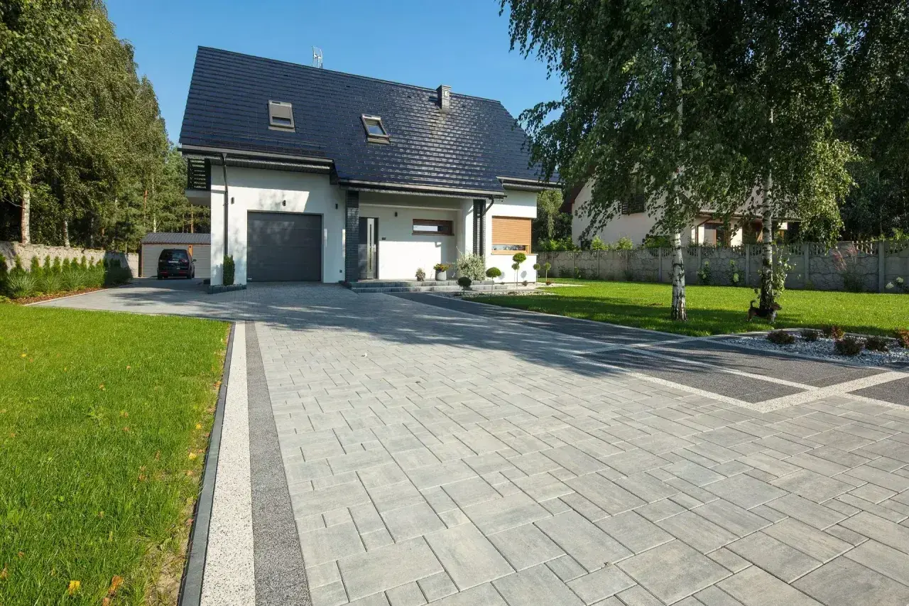Nowoczesny podjazd przed domem z szarej kostki brukowej, otoczony zielonym trawnikiem i drzewami.