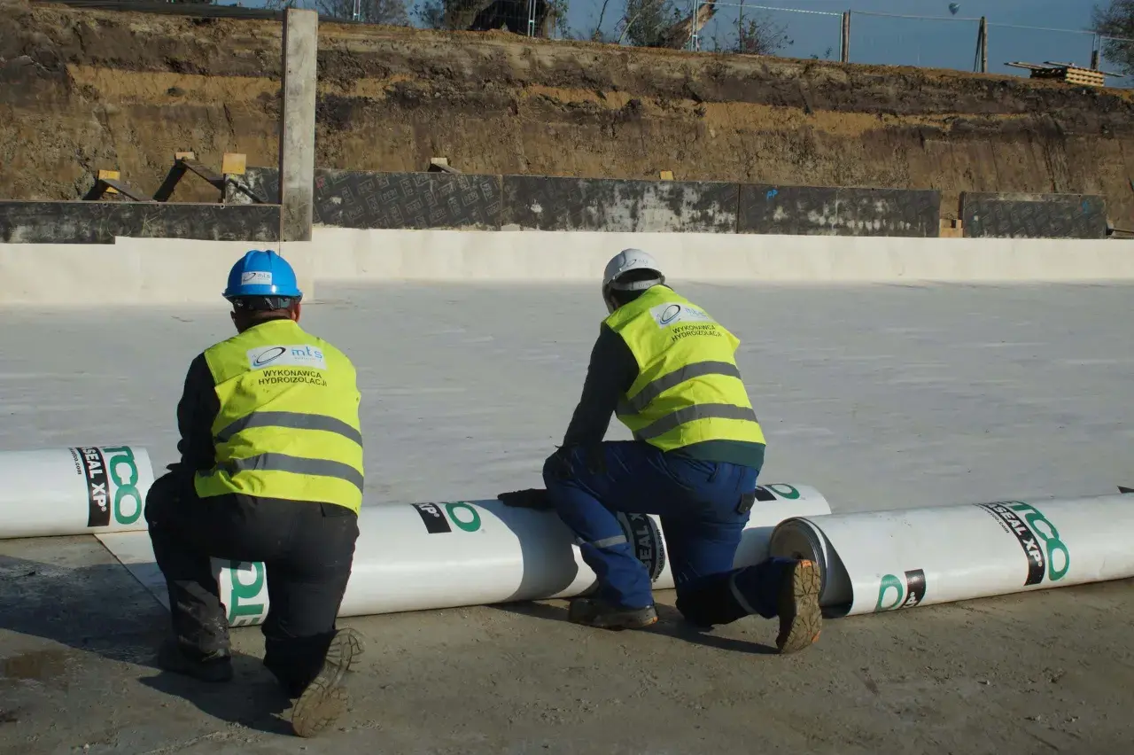 Dwóch pracowników w kamizelkach odblaskowych przygotowuje hydroizolację. Rozwijają rolki materiału, by zabezpieczyć teren budowy przed wilgocią.