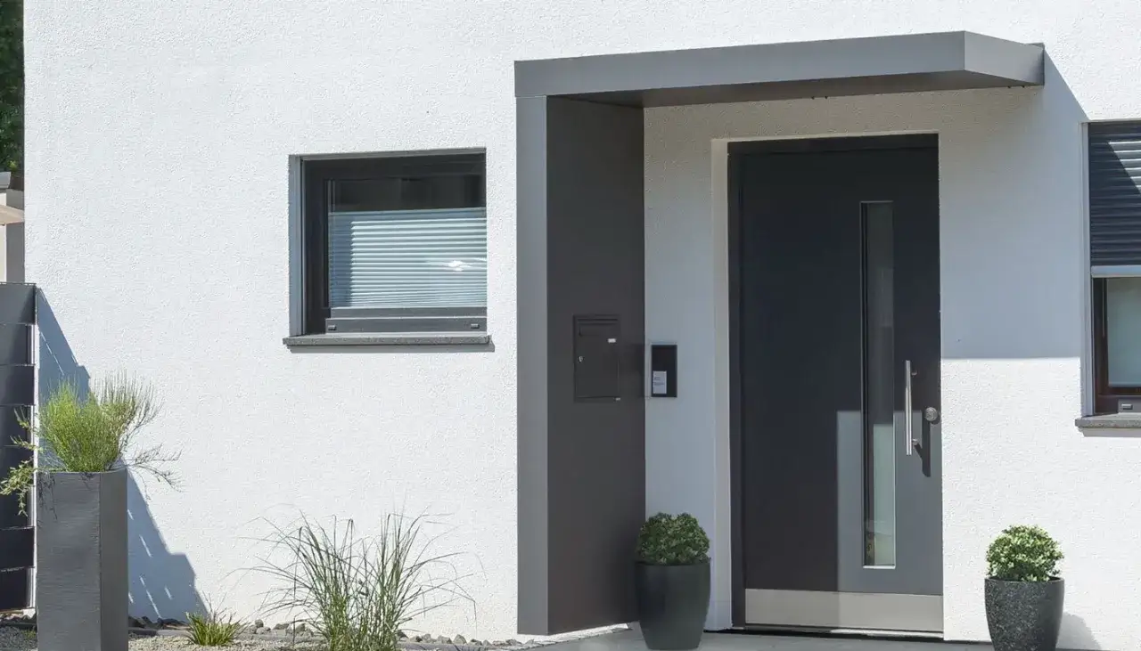 Nowoczesny projekt daszku nad drzwiami wejściowymi, z szarą, minimalistyczną formą, idealnie komponuje się z białym tynkiem i ciemnymi detalami.