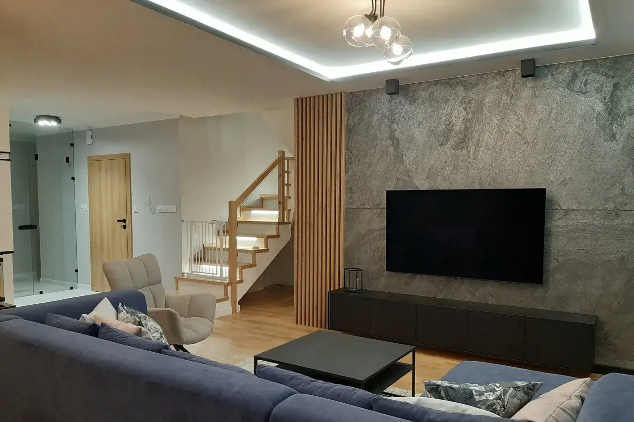 Nowoczesny salon z szarą ścianą, telewizorem i kanapą. Obok schodów pionowe rodzaje lameli drewnianych tworzą ciekawy akcent.