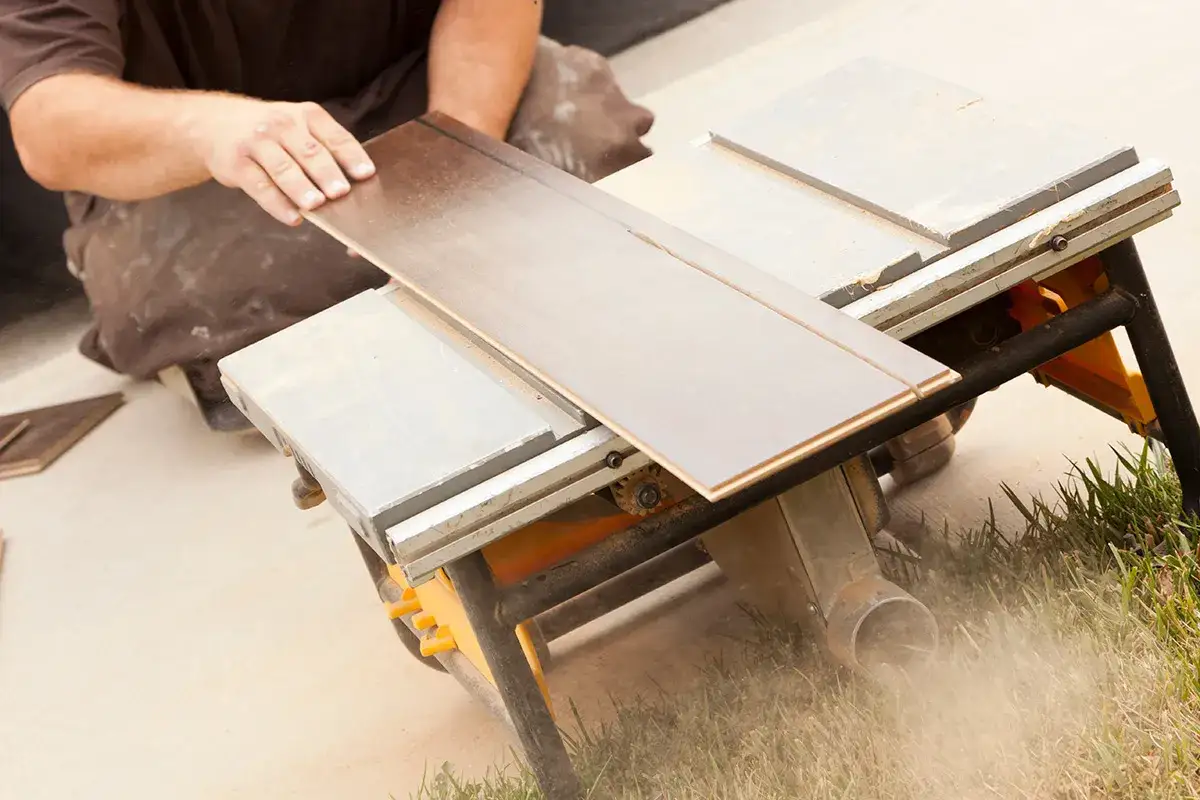 Ręce układają panel laminowany na pile stołowej, pokazując, czym ciąć panele laminowane. Pył unosi się w powietrzu.