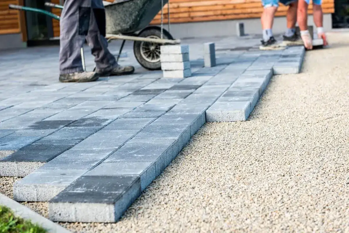 Układanie szarej kostki brukowej, która stanowi tańszy zamiennik kostki brukowej, na podsypce żwirowej. W tle widać taczkę i budynek.